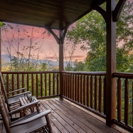 A covered balcony framed by rustic log railings opens to a stunning mountain view bathed in soft sunset hues, with rocking chairs waiting for quiet moments above the treetops.