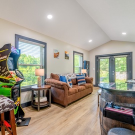 Kick back in your private game room featuring vintage arcade fun, a cozy couch for lounging, and doors opening to wooded views. The space is perfect for entertainment and relaxation during your stay.