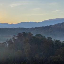 Morning mist drifts through layered mountain ridges as the sun rises over a peaceful forested landscape, painting the sky in soft pastels.