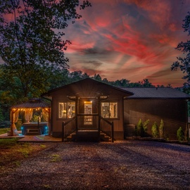 The property sits nestled among trees beneath a stunning sunset sky, with a welcoming exterior that glows warmly at dusk and a hot tub ready for relaxing evenings.
