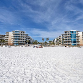 Beachfront property buildings overlook pristine white sand and turquoise waters under brilliant blue skies.
