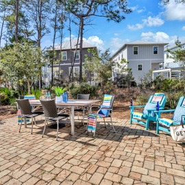 Your private brick patio beckons with colorful striped chairs and dining table, surrounded by towering pines and peaceful neighborhood views.