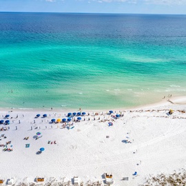 Pristine white sand beach stretches along crystal-clear turquoise waters with beachgoers enjoying the stunning Gulf Coast scenery.