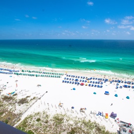 Stunning white sand beach with turquoise waters stretches endlessly below, featuring rows of blue umbrellas and beachgoers enjoying the pristine coastline.