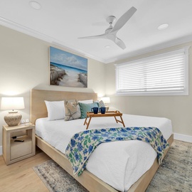 Relax in your coastal-inspired bedroom with plush bedding, a gentle ceiling fan, and peaceful beach artwork creating the perfect retreat.