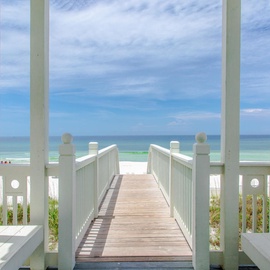Wooden boardwalk leads directly to pristine white sand beach with turquoise waters under partly cloudy skies.