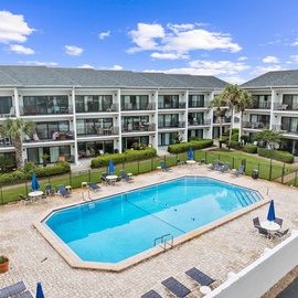 Multi-story resort buildings surrounding a sparkling central swimming pool with blue umbrellas and lounge chairs.