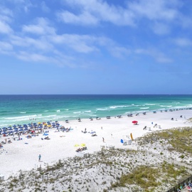 Pristine white sand beach stretches along turquoise waters, with colorful umbrellas dotting the shoreline under brilliant blue skies.
