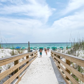 A wooden boardwalk leads through pristine dunes to a beautiful white sand beach with turquoise waters and colorful umbrellas.