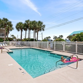 Community pool area surrounded by tropical palms and nearby buildings.