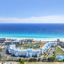 Stunning aerial perspective showcasing the beachfront property's prime coastal location, set against pristine emerald waters and sandy shores, surrounded by resort amenities including pools and tennis courts.