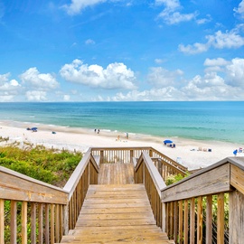 Wooden boardwalk leads to pristine white sand beach with turquoise waters and scattered beachgoers under a brilliant blue sky.