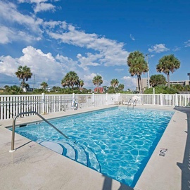 Emerald Towers West's sparkling swimming pool area features crystal-clear water surrounded by white safety fencing, with tropical palm trees and coastal buildings visible in the background under a beautiful blue sky.