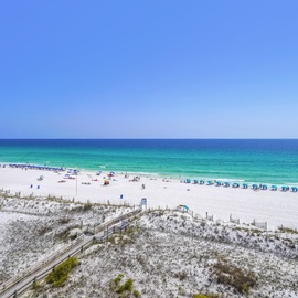 Pristine white sand beach with crystal-clear emerald waters, wooden boardwalk, and vibrant blue umbrals dotting the shoreline.