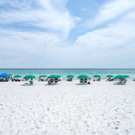 Pristine white sand beach stretches along turquoise waters with colorful umbrellas and chairs dotting the shoreline under blue skies.