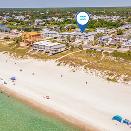 An aerial view showing the beachfront neighborhood and surrounding residential area. The property is located steps from sandy shores with calm, turquoise waters.