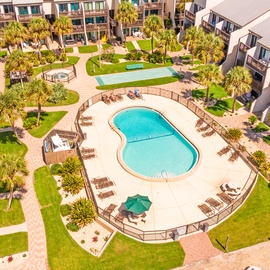 A bird's-eye view showcases the property's well-maintained pool area with surrounding sun loungers, lush green lawns, palm trees, and multi-story residential buildings arranged around a central courtyard.