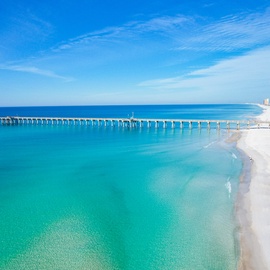 Stunning aerial view of pristine white sand beach with turquoise waters and fishing pier stretching into the Gulf.