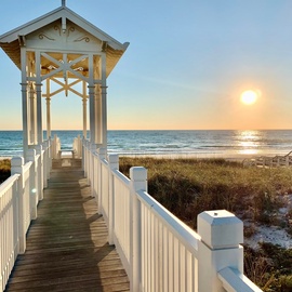 A charming white gazebo walkway leads to pristine beach views with golden sunset painting the ocean horizon.