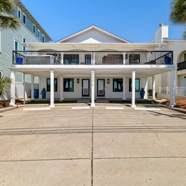 A coastal-style vacation rental nestled among palm trees in a residential beach community, featuring a covered porch and spacious second-floor balcony.