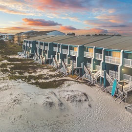 Beachfront vacation rentals with colorful exteriors line the pristine white sand dunes under a vibrant sunset sky.