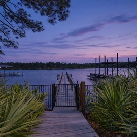 Tranquil lakefront dock at sunset, framed by native palms and towering pines, offering serene water views and private boat access at this peaceful waterfront property.