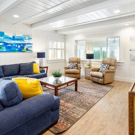 Relax in your bright, coastal-inspired living room with comfortable seating and vibrant ocean artwork adorning the white-paneled walls.