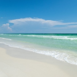 Pristine white sand beach with crystal-clear turquoise waters and gentle waves under a brilliant blue sky with scattered clouds.
