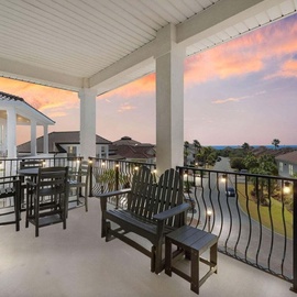 A covered balcony with dining and lounge seating overlooks a residential community beneath a vibrant sunset sky. The space provides a peaceful vantage point for evening gatherings.