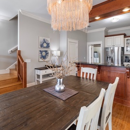 Beautiful wood dining table beneath a stunning statement chandelier. Open-concept space flows into a fully equipped kitchen with stainless steel appliances, warm wood cabinetry, and plenty of counter space.