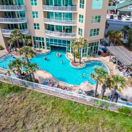 Aerial view of a beachfront resort featuring a curved swimming pool with tropical landscaping and multi-story accommodations.