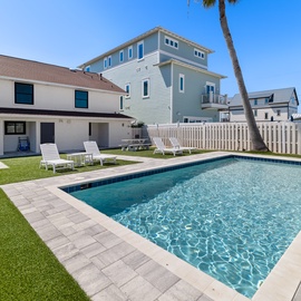 Your private backyard paradise awaits, with a sparkling pool, sun loungers, and lush green lawn surrounded by swaying palms and coastal charm.