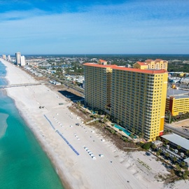 Beachfront resort towers rise above pristine white sand and emerald Gulf waters, with entertainment district nearby.