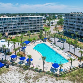Aerial view of a modern resort complex featuring a sparkling swimming pool surrounded by palm trees and blue umbrellas.
