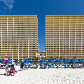 Beachfront twin towers rise majestically above pristine white sand, with blue umbrellas dotting the shoreline below.