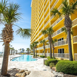 Bright yellow beachfront property featuring a sparkling pool area surrounded by swaying palm trees and manicured landscaping.