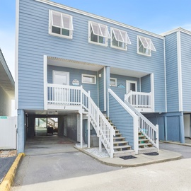 Charming blue coastal building with white railings and covered parking beneath your elevated unit.