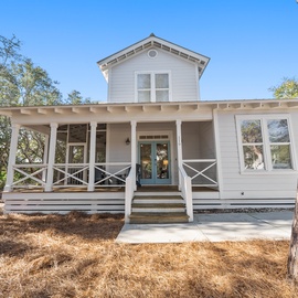 Charming white cottage with welcoming front porch and traditional farmhouse details, nestled among mature trees in a peaceful setting.