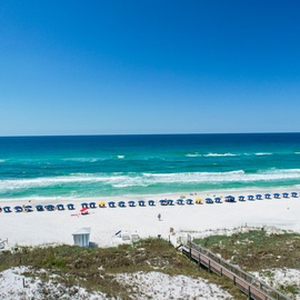 Pristine white sand beach with turquoise waters and organized beach umbrellas stretches along the coastline under clear blue skies.