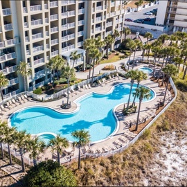 Aerial view of beachside resort featuring curved swimming pools surrounded by palm trees and multi-story buildings with balconies.