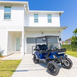 Pristine two-story coastal retreat with convenient golf cart parking and lush tropical landscaping beneath a brilliant blue sky.