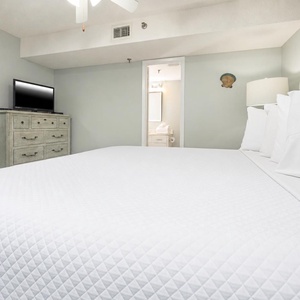 The bedroom features a large king bed with white linens, a wooden dresser, and a TV, with an en-suite bathroom