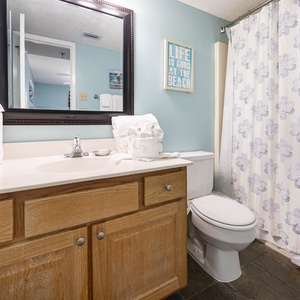 The geust bath features a light blue wall, a wooden vanity with a sink, and a floral shower curtain, with a refreshing vibe