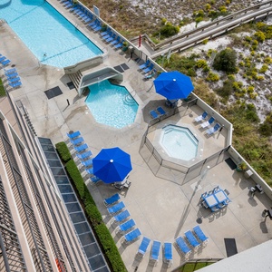 Aerial view of the pool area featuring a large pool, kiddie pool, blue umbrellas, and ample seating for relaxation