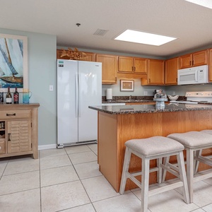 Cozy kitchen area features light wood cabinetry, a breakfast bar with three stools, and a decorative coastal-themed artwork