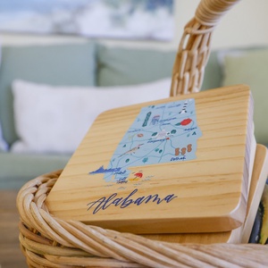 Decorative wooden coasters featuring a colorful map of Alabama sit in a woven basket enhance that coastal feel