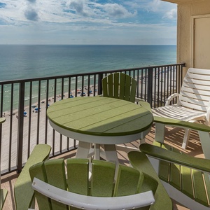 The balcony features green and white outdoor seating with a view of the beach and umbrellas lining the shore below