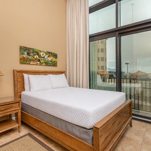 This bedroom features a cozy queen bed, a stylish lamp, and large windows offering a bright view of the surrounding area