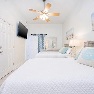 Bright guest bedroom featuring two double beds, a wall-mounted TV, and coastal decor that enhances a relaxing beach vibe