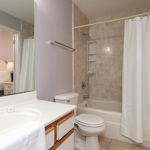 Bright bathroom featuring a tub/shower combo, single sink vanity, and a glimpse of a cozy guest bedroom beyond the door